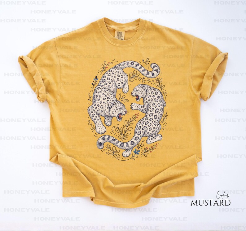 May include: A mustard yellow short-sleeve t-shirt featuring a central graphic of two snow leopards in a circular arrangement surrounded by botanical line art. The leopards are depicted in white with black spots, facing each other in a dynamic pose.