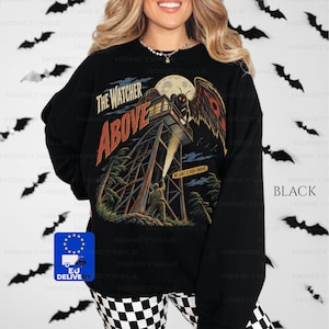 May include: A black long-sleeve sweatshirt featuring a graphic print of a fire lookout tower under a full moon with a moth-like creature at the top. The text reads THE WATCHER ABOVE and NO LIGHT IS HIGH ENOUGH. The design includes trees and a person.