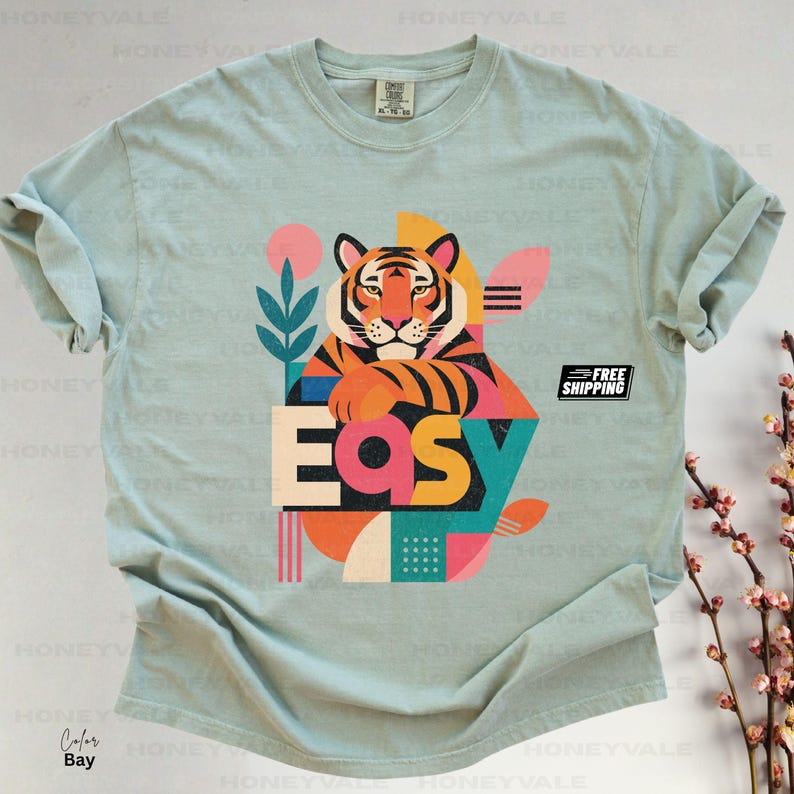 Pode incluir: Uma t-shirt de manga curta em verde s&aacute;lvia claro com uma ilustra&ccedil;&atilde;o gr&aacute;fica de um tigre com riscas cor de laranja, pretas e brancas. O design inclui a palavra "EASY" em letras grandes e coloridas, juntamente com formas geom&eacute;tricas e motivos vegetais.