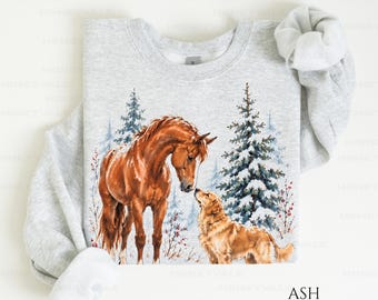 May include: A light gray crewneck sweatshirt featuring a graphic illustration of a brown horse and a golden retriever dog standing together in a snowy forest with evergreen trees and red berries. The word ASH is printed in the bottom right corner.