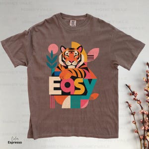 Pode incluir: Uma t-shirt de manga curta cor de caf&eacute; expresso com uma impress&atilde;o gr&aacute;fica de um tigre a apoiar as patas na palavra "Easy". O design incorpora formas geom&eacute;tricas e elementos bot&acirc;nicos em tons de rosa, azul-petr&oacute;leo, laranja e amarelo.