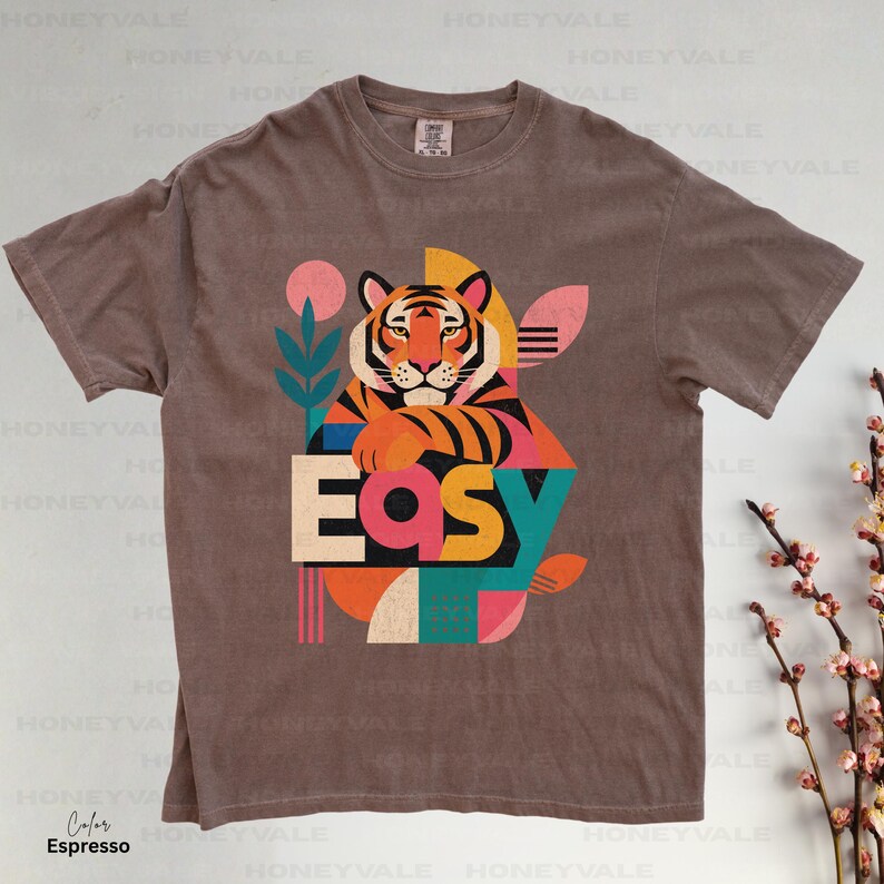 Pode incluir: Uma t-shirt de manga curta cor de caf&eacute; expresso com uma impress&atilde;o gr&aacute;fica de um tigre a apoiar as patas na palavra "Easy". O design incorpora formas geom&eacute;tricas e elementos bot&acirc;nicos em tons de rosa, azul-petr&oacute;leo, laranja e amarelo.