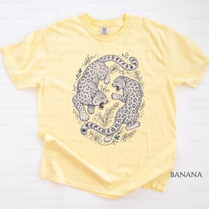 May include: A pale yellow short-sleeve t-shirt featuring a central graphic of two snow leopards in a circular arrangement surrounded by botanical line art. The leopards are depicted in shades of grey and white with black spots and open mouths.