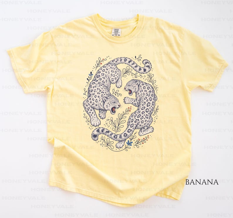 May include: A pale yellow short-sleeve t-shirt featuring a central graphic of two snow leopards in a circular arrangement surrounded by botanical line art. The leopards are depicted in shades of grey and white with black spots and open mouths.