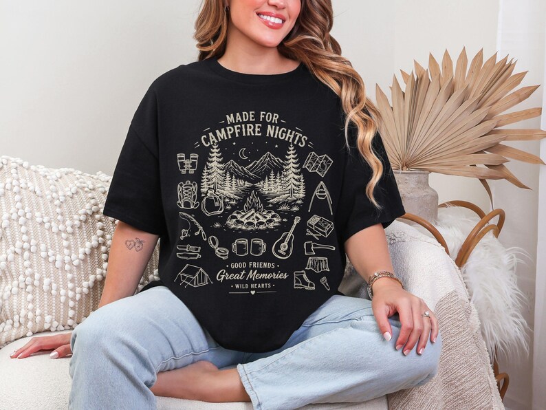 May include: A black short-sleeve t-shirt featuring a cream-colored graphic of a campfire scene with mountains, trees, and camping gear. The text reads: MADE FOR CAMPFIRE NIGHTS, GOOD FRIENDS, Great Memories, WILD HEARTS.