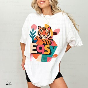 Pode incluir: Uma t-shirt branca oversize com uma impress&atilde;o gr&aacute;fica de um tigre com riscas cor de laranja e pretas. O design inclui a palavra "EASY" em letras de bloco arrojadas e coloridas, rodeada por formas abstratas em tons de rosa, azul-petr&oacute;leo e amarelo.