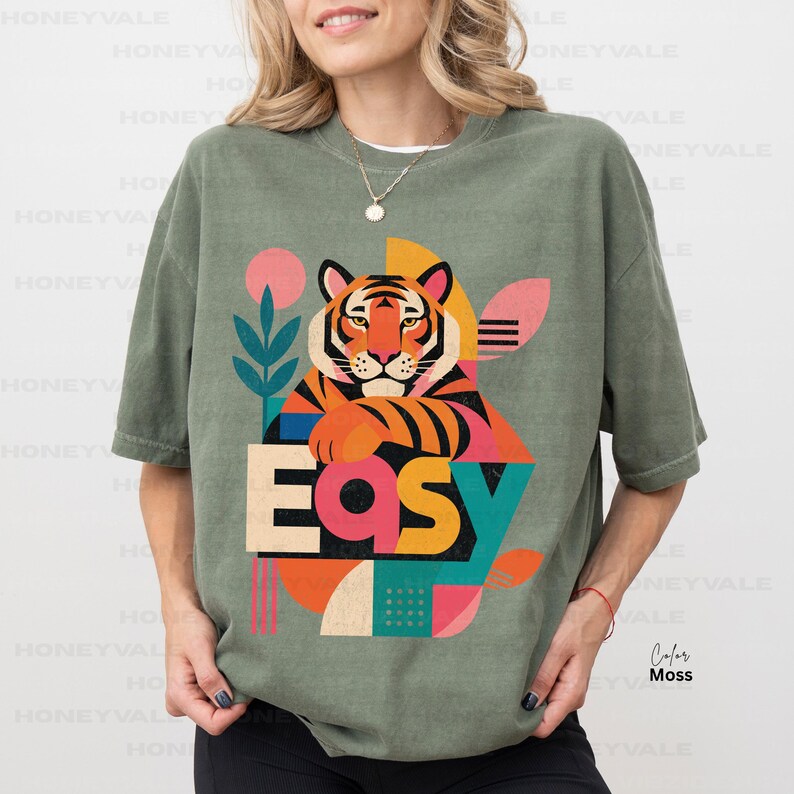 Pode incluir: Uma t-shirt de manga curta verde-azeitona com uma impress&atilde;o gr&aacute;fica de um tigre e a palavra "EASY" em letras grandes e coloridas. O design inclui formas geom&eacute;tricas em rosa, laranja, amarelo e turquesa, juntamente com um motivo vegetal.