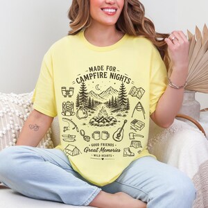 May include: A pale yellow short-sleeve t-shirt featuring a black graphic print of a campfire scene with mountains, trees, and camping gear. The text reads MADE FOR CAMPFIRE NIGHTS, GOOD FRIENDS, GREAT MEMORIES, and WILD HEARTS in a decorative font.