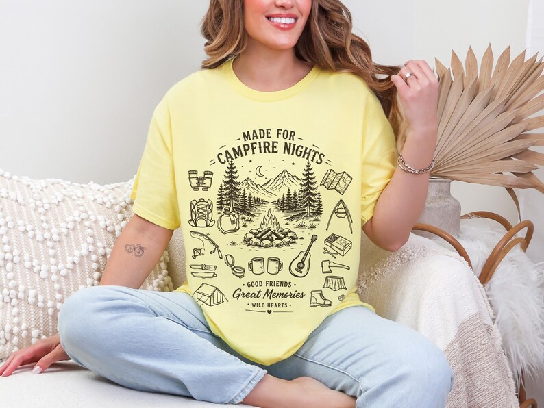May include: A pale yellow short-sleeve t-shirt featuring a black graphic print of a campfire scene with mountains, trees, and camping gear. The text reads MADE FOR CAMPFIRE NIGHTS, GOOD FRIENDS, GREAT MEMORIES, and WILD HEARTS in a decorative font.