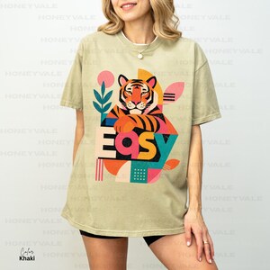 Pode incluir: Uma t-shirt de manga curta cor de c&aacute;qui com uma estampa gr&aacute;fica de um rosto de tigre acima da palavra EASY em letras grandes e coloridas. O design inclui formas abstratas em rosa, laranja, azul-petr&oacute;leo e amarelo, com uma planta.