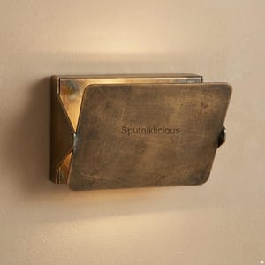 Könnte beinhalten: Eine rechteckige Wandleuchte aus gealtertem Messing mit verwitterter Metalloberfläche. Auf der Vorderseite ist das Wort Sputniklicious eingraviert. Das Design verfügt über seitliche Scharniere, die Licht nach oben austreten lassen.
