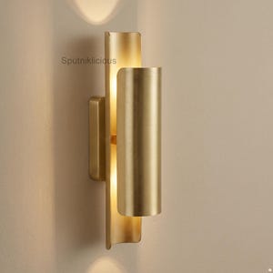 Könnte beinhalten: Eine Wandleuchte aus gebürstetem Gold mit zwei vertikalen, geschwungenen Metallzylindern auf einem rechteckigen Sockel. Die Leuchte strahlt ein warmes, sanftes Licht hinter den Zylindern aus und erzeugt Lichtmuster an der Wand. Der Text Sputniklicious ist sichtbar.