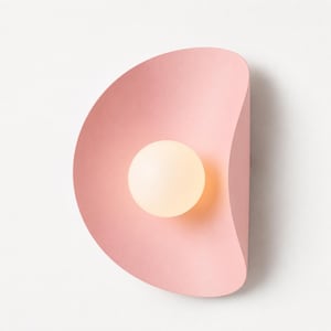 May include: A wall sconce featuring a curved, shell-like pink metal shade with a smooth matte finish. Centered within the concave curve of the shade is a single, spherical, frosted white glass light bulb that emits a soft, warm glow.