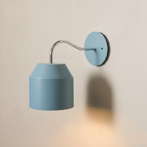 May include: A light blue wall sconce with a cylindrical shade and a curved, silver-toned arm. The sconce has a circular mounting plate and is designed for indoor lighting. The fixture is a modern design, suitable for bedrooms, hallways, or living rooms.