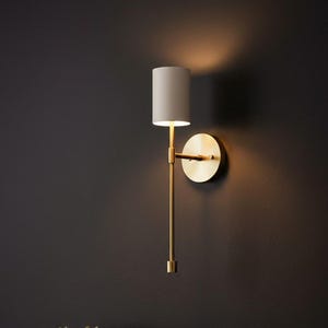 Peut inclure: Une applique murale composée d'une fine tige métallique verticale de couleur laiton et d'une plaque murale circulaire en laiton. Le luminaire est surmonté d'un abat-jour cylindrique en tissu blanc cassé diffusant une lumière chaude.