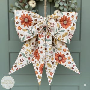 May include: A large, decorative bow with a fall floral pattern. The bow features orange flowers, green leaves, and acorns on a cream-colored background. It is suspended by a natural twine string, perfect for seasonal decor.