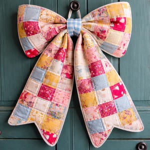 Op de afbeelding: Een grote, decoratieve strik gemaakt van patchworkstof in roze, gele en blauwe tinten. De strik is gewatteerd en heeft een ruitjesmidden. Hij hangt aan een turquoise deur.