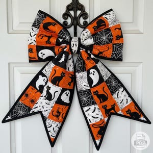 May include: A large Halloween-themed bow with a patchwork design. The bow features orange, black, and white squares with images of black cats, ghosts, spiders, and spiderwebs. The bow is trimmed with black fabric and is hanging on a white door.