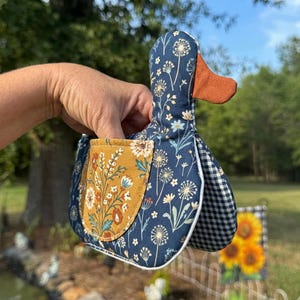 May include: A fabric duck-shaped oven mitt featuring a dark blue background with a white dandelion print, a solid rust-colored beak, and a mustard yellow pocket with a floral pattern. The side panel displays a black and white gingham check.