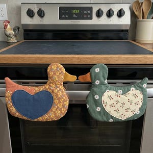 May include: Two fabric oven mitts shaped like ducks hanging on an oven handle. The left duck has a yellow floral pattern with a solid blue heart patch. The right duck is green with a white daisy and red berry pattern and a heart patch.
