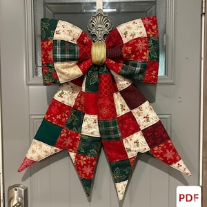 May include: A large, quilted bow in a patchwork design, featuring red, green, and cream fabrics with floral and plaid patterns. The bow is adorned with a gold band in the center and a decorative silver accent. It is hanging on a gray door.