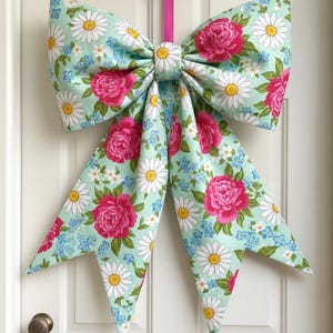 May include: A large fabric bow with a floral pattern of pink roses, white daisies, and blue forget-me-nots on a light teal background. The bow is hanging on a white door with a gold-colored hook and a pink ribbon.