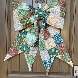 May include: A large, decorative bow made of patchwork fabric, featuring various patterns including floral, plaid, and fox motifs. The bow is predominantly green, brown, and orange, and is designed to be hung on a door.