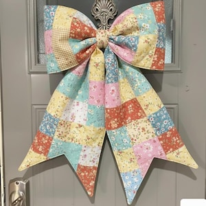 May include: A large, quilted fabric bow in a patchwork design, featuring squares of floral and patterned fabrics in pastel colors. The bow is attached to a door with a decorative metal fixture. The bow's colors include pink, yellow, blue, and orange.