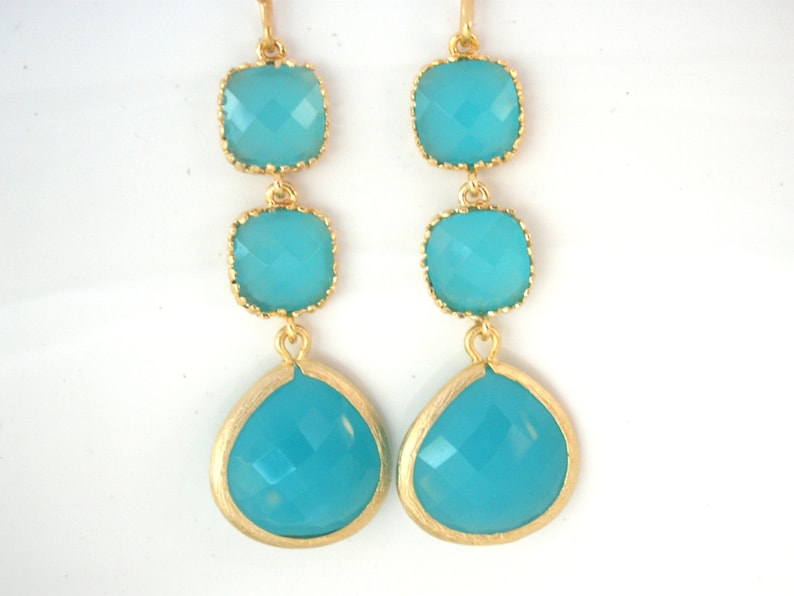 May include: Gold earrings with three turquoise blue gemstone drops. The top two gemstones are square and the bottom gemstone is teardrop shaped.