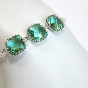 Può includere: Un braccialetto a catena d'argento con tre pietre preziose quadrate di colore verde chiaro. Ogni pietra preziosa è circondata da un bordo argentato.