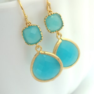 May include: Gold dangle earrings with two turquoise blue gemstone drops. The top gemstone is square and the bottom gemstone is teardrop shaped.
