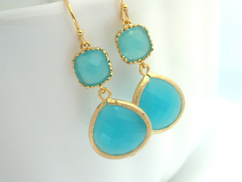 May include: Gold dangle earrings with two turquoise blue gemstone drops. The top gemstone is square and the bottom gemstone is teardrop shaped.