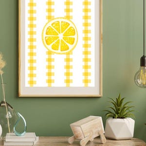 May include: Framed art print featuring a yellow lemon slice on a white and yellow gingham pattern background. The artwork is displayed in a light brown wooden frame, suitable for kitchen or dining room decor.