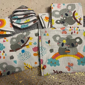 May include: A collection of baby items featuring a koala bear theme. The set includes several gray washcloths, reusable wipes, and fabric squares with koala illustrations, rainbows, and colorful accents. The items are arranged on a textured surface.