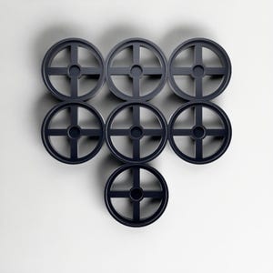 May include: Seven dark grey, circular objects with a cross-shaped design are arranged in a pattern against a white background. The objects appear to be made of a solid material and have a minimalist aesthetic.