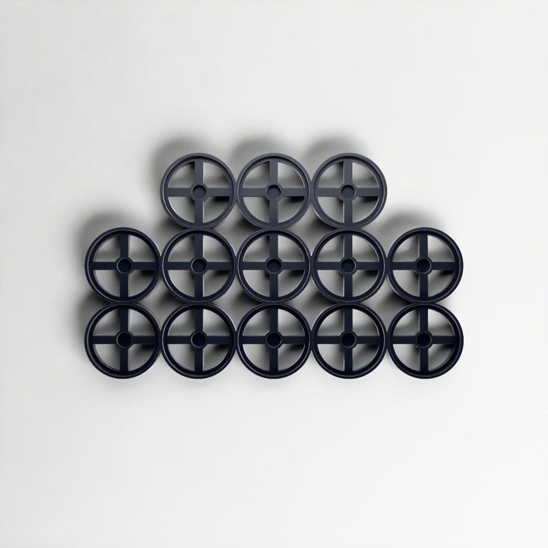 May include: Twelve dark grey plastic wheels with a cross-shaped design are arranged in a grid pattern. The wheels are evenly spaced against a white background. These wheels are likely for a toy or model.