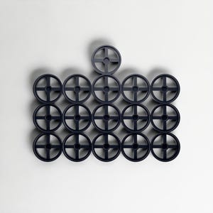 May include: A collection of dark grey circular objects, each featuring a cross-shaped design at the centre. They are arranged in a grid formation against a white backdrop, forming a geometric pattern.