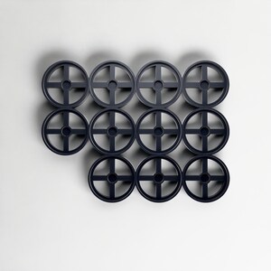 May include: Twelve dark grey circular objects, each with a cross-shaped design and a central hole, arranged against a white background. The objects are evenly spaced and appear to be made of plastic or a similar material.