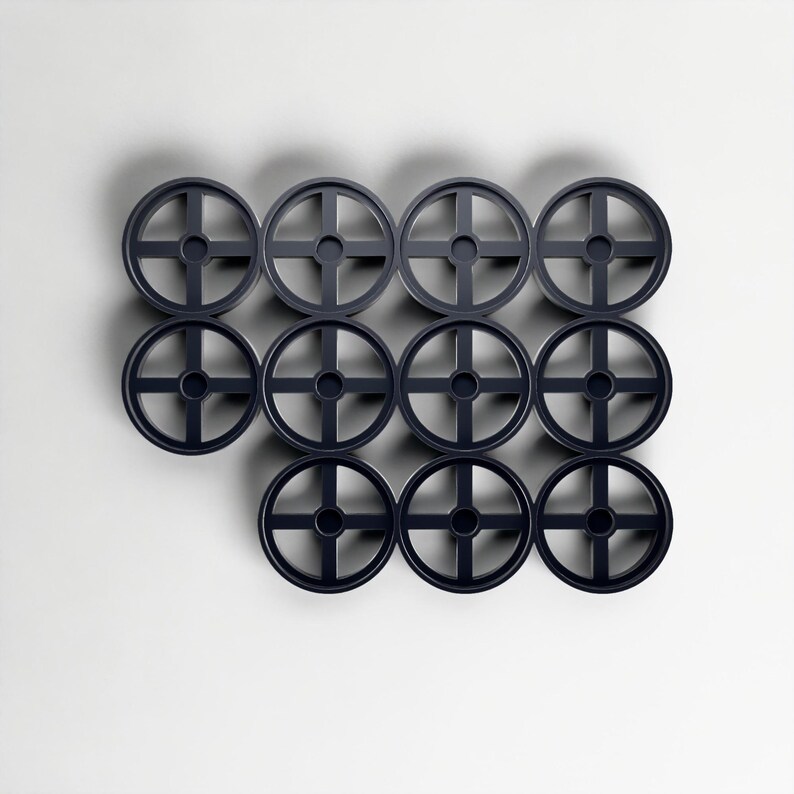 May include: Twelve dark grey circular objects, each with a cross-shaped design and a central hole, arranged against a white background. The objects are evenly spaced and appear to be made of plastic or a similar material.