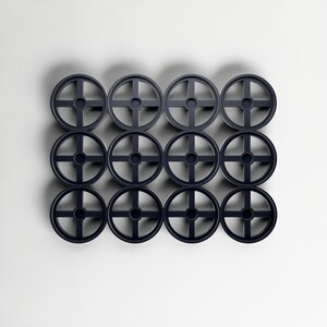 May include: A grid of twelve dark grey circular objects, each with a cross-shaped centre and a wheel-like design. The objects are arranged in a neat pattern against a plain white background. Each object has a diameter of approximately 10 cm.