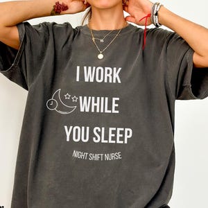 Puede incluir: Camiseta gris oscuro con texto blanco: "I WORK WHILE YOU SLEEP NIGHT SHIFT NURSE". El diseño incluye una luna creciente, estrellas y un reloj. La camiseta es de corte holgado.