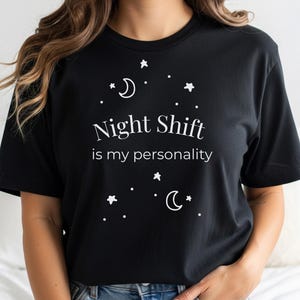 Puede incluir: Camiseta negra con el texto blanco "Night Shift is my personality", junto con ilustraciones blancas de estrellas y lunas crecientes. La camiseta es de cuello redondo.