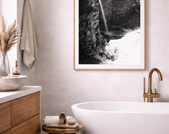 May include: A black and white framed print of a waterfall cascading down a rocky cliff, hanging above a white bathtub. The bathroom features a wooden vanity, a small wooden stool, and a brass faucet.