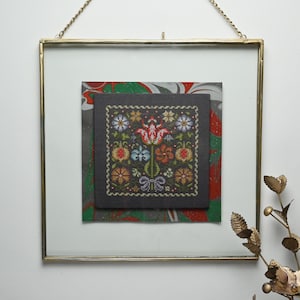 May include: A framed cross-stitch floral design. The artwork features a dark background with a colourful flower pattern. The frame is gold-toned, and the artwork is displayed with a white mount and a decorative paper background.