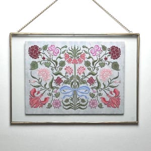 May include: A framed cross-stitch with a floral pattern in pink, red, and green. The embroidery depicts flowers, leaves, and a blue bow. The gold-coloured frame is suspended by a chain.