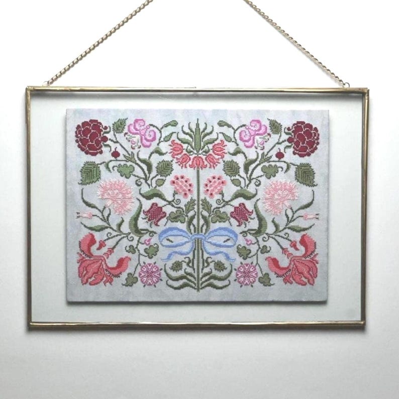 May include: A framed cross-stitch with a floral pattern in pink, red, and green. The embroidery depicts flowers, leaves, and a blue bow. The gold-coloured frame is suspended by a chain.