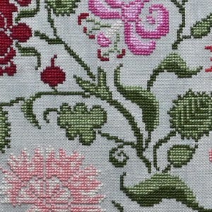 May include: Close-up of a cross-stitch floral pattern. The design features red, pink, and green flowers and leaves on a light grey background. The needlework is detailed, showcasing the texture of the stitches.