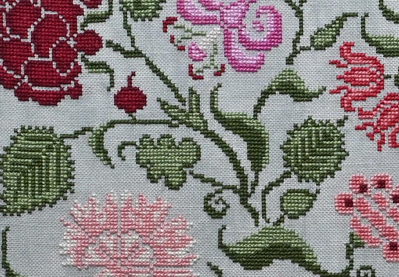 May include: Close-up of a cross-stitch floral pattern. The design features red, pink, and green flowers and leaves on a light grey background. The needlework is detailed, showcasing the texture of the stitches.