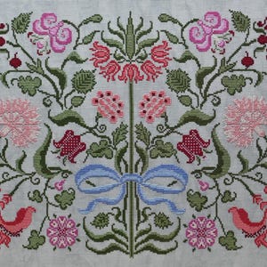 May include: Cross-stitch floral design with pink, red, and green flowers and foliage on a light grey background. A blue ribbon is centered at the bottom. The design includes various flower types and detailed stitching.