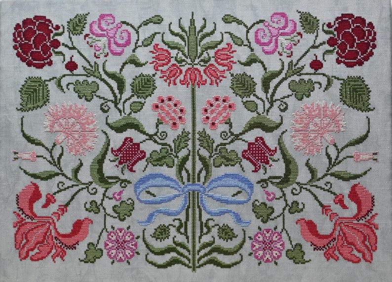 May include: Cross-stitch floral design with pink, red, and green flowers and foliage on a light grey background. A blue ribbon is centered at the bottom. The design includes various flower types and detailed stitching.